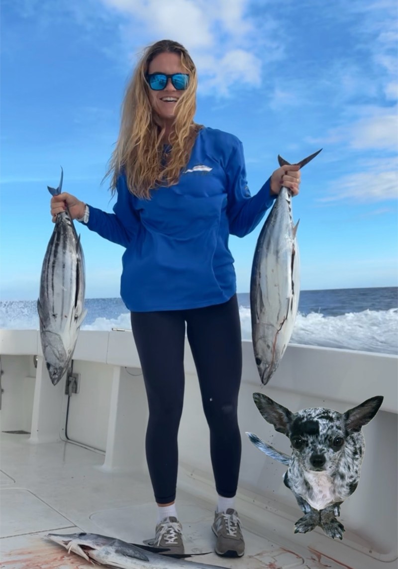 Person in blue shirt holding two fish on a boat with a small dog nearby.