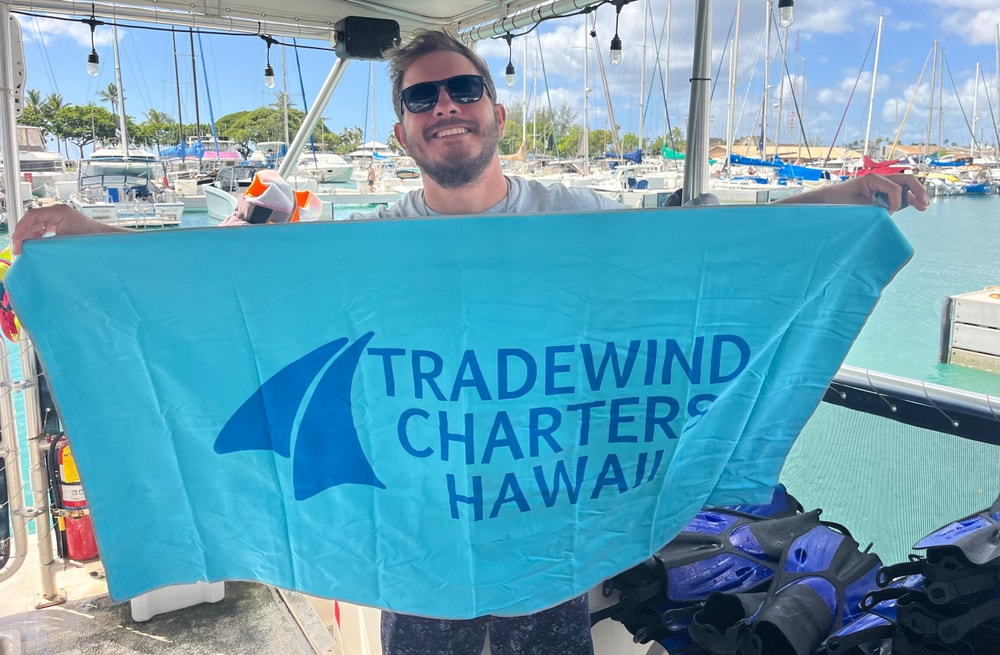 Person holding a blue 'Tradewind Charters Hawaii' towel on a boat with harbored yachts in the background.