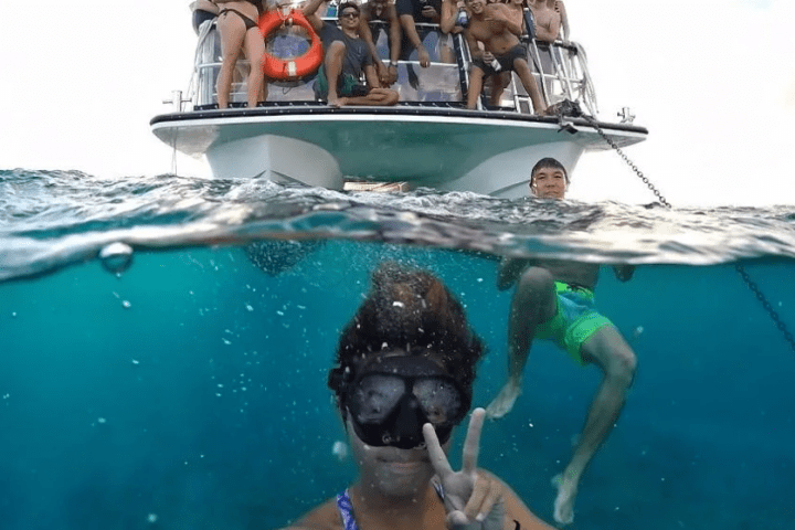 Group of people on a boat, one person underwater taking a selfie, another in water, ocean view.