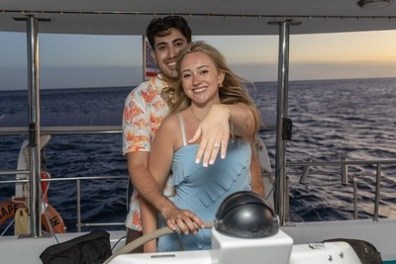 Couple on a boat, woman shows engagement ring at sunset.
