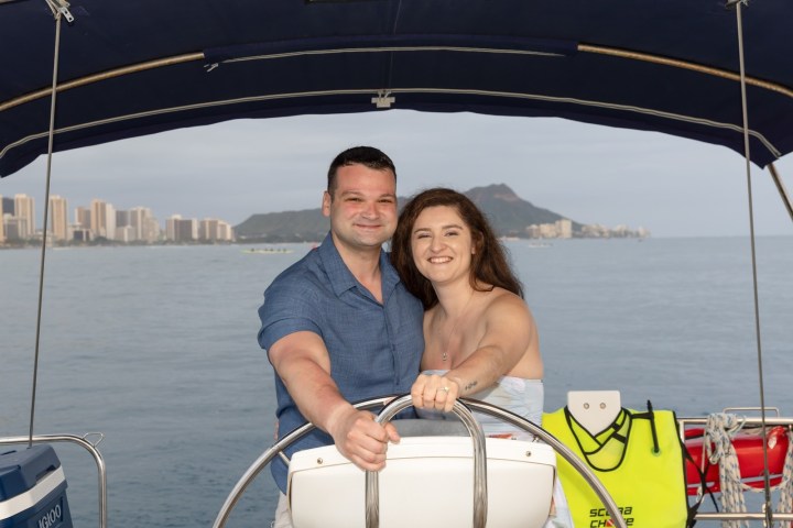 Couple smiling on a boat with cityscape and mountain in background.