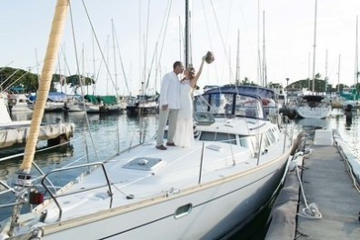 Couple in white attire celebrating on a docked sailboat at a marina.